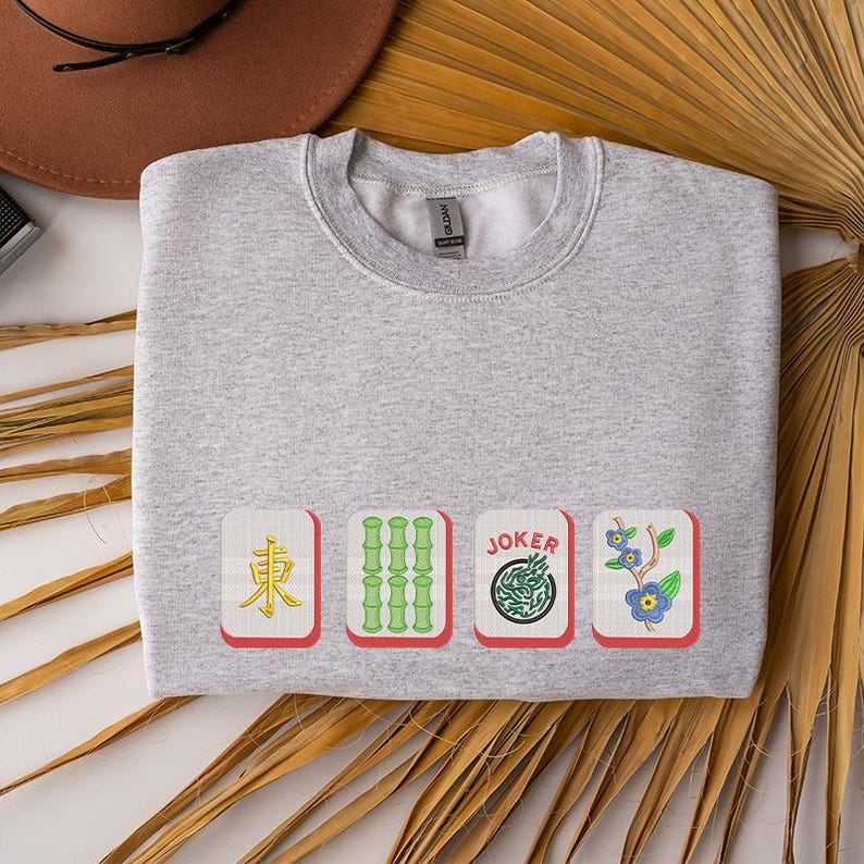 May include: A light gray sweatshirt featuring four Mahjong tile designs. The tiles include a red East wind tile, green bamboo, a Joker tile, and a blue flower tile. The sweatshirt is folded on a white surface.