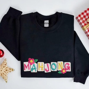 Puede incluir: Sudadera negra con la palabra "MAHJONG" bordada en letras coloridas, adornada con flores rosas y hojas verdes. Una caja de regalo y un adorno en forma de estrella son visibles en el fondo.