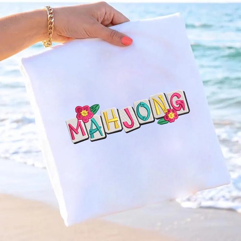 Puede incluir: Coj&iacute;n cuadrado blanco con la palabra "MAHJONG" bordada en letras coloridas. Las letras est&aacute;n delineadas en negro y adornadas con flores rosas y hojas verdes. El coj&iacute;n es sostenido por una persona con una pulsera dorada.