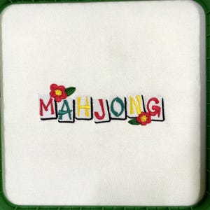 Puede incluir: Tela blanca con la palabra "MAHJONG" bordada en letras de bloque coloridas. Flores rojas y amarillas con hojas verdes est&aacute;n bordadas cerca de las letras. La tela est&aacute; dentro de un marco cuadrado verde.