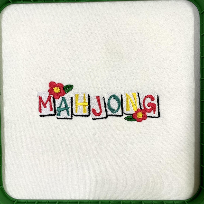 Puede incluir: Tela blanca con la palabra "MAHJONG" bordada en letras de bloque coloridas. Flores rojas y amarillas con hojas verdes est&aacute;n bordadas cerca de las letras. La tela est&aacute; dentro de un marco cuadrado verde.