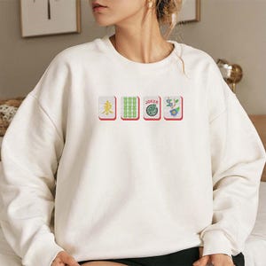 May include: White sweatshirt featuring four embroidered Mahjong tiles. The tiles include a red-bordered tile with a yellow character, a green bamboo tile, a tile with the word "Joker", and a floral tile.