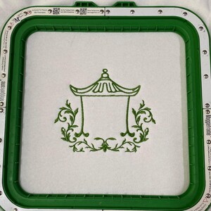 May include: A green embroidery hoop frame holding white fabric with a centered green stitched design of a pagoda structure surrounded by ornate leafy scrollwork. The frame features metallic silver accents and printed text along the edges.