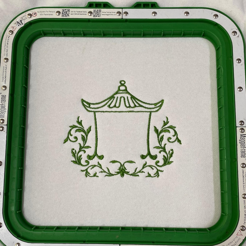 May include: A green embroidery hoop frame holding white fabric with a centered green stitched design of a pagoda structure surrounded by ornate leafy scrollwork. The frame features metallic silver accents and printed text along the edges.