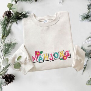Puede incluir: Sudadera color crema con la palabra "MAHJONG" bordada en letras coloridas y detalles florales. La sudadera tiene cuello redondo y mangas largas. El dise&ntilde;o est&aacute; centrado en la parte delantera.