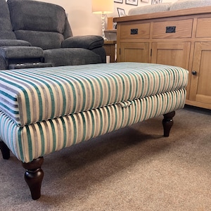 May include: A striped ottoman in teal, white, grey, and beige, with turned wooden legs. The rectangular upholstered footstool is placed on a carpet. The background includes a grey sofa and a wooden cabinet. The ottoman's length is approximately 120 cm.