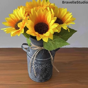 May include: A bouquet of bright yellow sunflowers with brown centers and green leaves in a distressed gray metal vase. The vase has a heart design and is tied with twine. The text "BravelleStudio" is in the upper right corner.