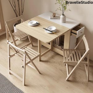May include: A light wood dining set with a folding table and three matching folding chairs. The table has a white top section and a drawer. Two white plates and chopsticks are on the table. A vase with greenery is on the table.