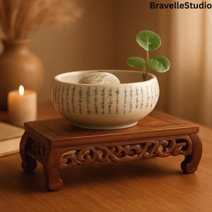 May include: A small, off-white ceramic bowl with black script and a stone inside, holding a small plant. The bowl sits on a dark brown, carved wooden stand. The image is set on a wooden surface.