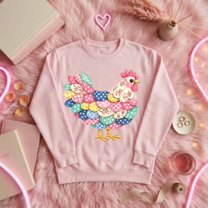 Puede incluir: Sudadera rosa claro con un diseño de gallina colorida. La gallina tiene un cuerpo con estampado floral, una cresta roja y un pico y patas amarillas. Las plumas de la cola son una mezcla de lunares y cuadros vichy en varios colores.