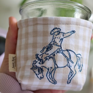 Könnte beinhalten: Ein klares Glas mit einer beige-weiß karierten Stoffhülle. Die Hülle hat ein blaues Stickdesign eines Cowboys auf einem Pferd. Das Etikett lautet "Josie Rae Handmade By".
