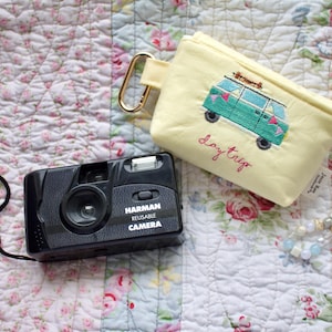 Può includere: Una fotocamera Harman riutilizzabile nera con cinturino da polso è accanto a una pochette giallo chiaro ricamata con un furgone turchese e le parole "Day Trip". È presente anche una collana di perline, il tutto su una trapunta floreale.