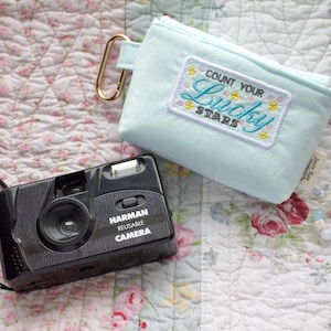May include: A black Harman reusable camera with a wrist strap sits next to a light blue pouch with a gold carabiner. The pouch has an embroidered patch that reads "Count Your Lucky Stars". A beaded necklace is also present.