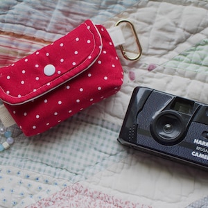 May include: A red fabric pouch with white polka dots, a white snap closure, and a gold-tone carabiner, accompanied by a beaded wrist strap and a black Harman Reusable Camera with a lens, flash, and white text on the front body.