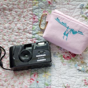 May include: A black Harman reusable camera with a black strap, a light pink pouch with "Sea, Salt, Sun" embroidered on it, and a beaded necklace with blue and white beads, all on a floral patterned quilt.
