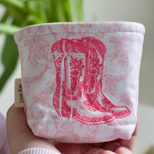Könnte beinhalten: Kleiner, zylindrischer Stoffbehälter mit rosa Blumenmuster und einem Paar gestickter rosa Cowboy-Stiefel. Das Etikett "Josie Rae" ist auf der Seite sichtbar. Der Behälter wird von einer Person in der Hand gehalten.