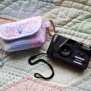 May include: A small fabric pouch with a pink floral and blue gingham pattern, featuring a blue snap closure and a gold carabiner. Attached is a beaded wrist strap with pastel beads. Next to it is a black Harman Reusable Camera with a black wrist strap.