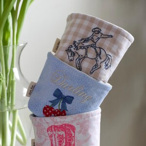 Peut inclure: Trois contenants en tissu empil&eacute;s. Le sup&eacute;rieur pr&eacute;sente un motif &agrave; carreaux beiges et blancs avec un cow-boy &agrave; cheval. Le milieu est bleu clair avec le mot "Darling" brod&eacute;, un n&oelig;ud et des cerises. Le bas a un motif floral rose avec une botte de cowboy rose.