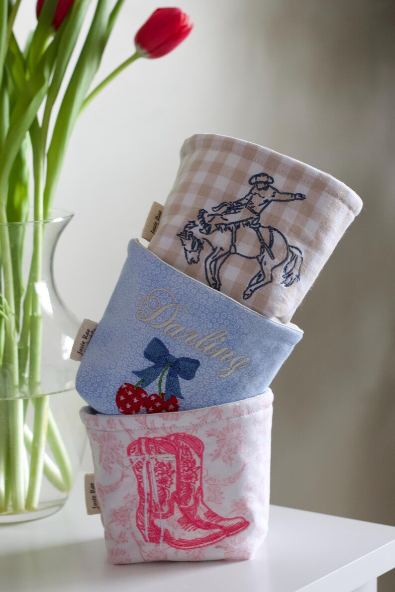 Peut inclure: Trois contenants en tissu empil&eacute;s. Le sup&eacute;rieur pr&eacute;sente un motif &agrave; carreaux beiges et blancs avec un cow-boy &agrave; cheval. Le milieu est bleu clair avec le mot "Darling" brod&eacute;, un n&oelig;ud et des cerises. Le bas a un motif floral rose avec une botte de cowboy rose.