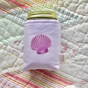 Peut inclure: Un étui de pot violet clair avec un couvercle doré, orné d'un motif de coquillage magenta brodé. L'étui est en tissu doux et est conçu pour s'adapter à un pot standard. Une petite étiquette est attachée sur le côté.