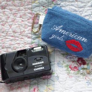 Puede incluir: Una cámara Harman reutilizable negra con una correa negra está junto a una bolsa de mezclilla bordada con "American girls" y un diseño de labios rojos. También se ve un cierre dorado y una pulsera de cuentas.