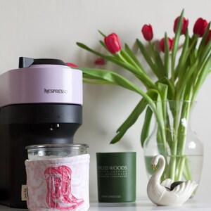 Peut inclure: Une cafeti&egrave;re Nespresso lavande et noire est pos&eacute;e &agrave; c&ocirc;t&eacute; d'un bocal en verre avec une housse rose sur le th&egrave;me des bottes. Une bougie verte et une figurine de cygne blanc sont &eacute;galement sur la surface blanche. Un vase de tulipes rouges compl&egrave;te la sc&egrave;ne.