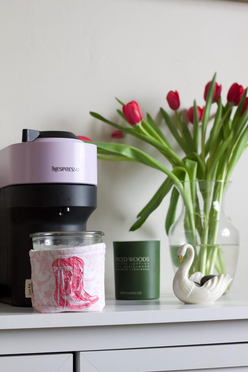 Peut inclure: Une cafeti&egrave;re Nespresso lavande et noire est pos&eacute;e &agrave; c&ocirc;t&eacute; d'un bocal en verre avec une housse rose sur le th&egrave;me des bottes. Une bougie verte et une figurine de cygne blanc sont &eacute;galement sur la surface blanche. Un vase de tulipes rouges compl&egrave;te la sc&egrave;ne.