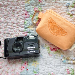 Może przedstawiać: Czarny, wielokrotnego użytku aparat Harman z czarnym paskiem, brzoskwiniowa torebka z haftowanym plasterkiem pomarańczy i naszyjnik z koralików z niebieskimi i białymi koralikami, wszystko na kwiecistej kołdrze.