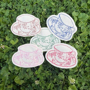 May include: Five white fabric patches shaped like tea cups and saucers, each featuring a different colored embroidered outline of a floral pattern. The colors of the embroidery are red, blue, green, pink, and a darker red, all on white fabric.