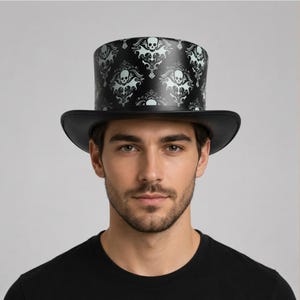 May include: A black top hat featuring a repeating white damask-style pattern of skulls and ornate scrollwork. The hat has a flat crown and a curved brim, with the graphic print covering the entire cylindrical side of the hat structure.