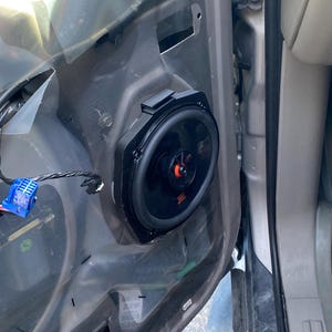 Puede incluir: Un altavoz de coche JBL negro instalado en la puerta de un vehículo. El altavoz es redondo con un marco negro y un centro rojo. Se ve el cableado y un conector azul.