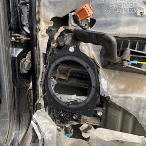 May include: An open car door panel with exposed components. A black speaker ring is visible, along with wiring, an orange connector, and a small motor. The panel is partially disassembled, revealing the interior structure.
