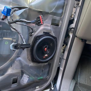 Pode incluir: Painel da porta do carro com um alto-falante JBL preto instalado. O alto-falante é montado em uma moldura preta. Chicotes de fios com conectores azuis e vermelhos são visíveis, juntamente com outros componentes.