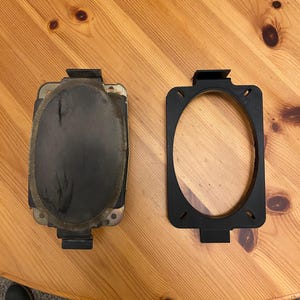 May include: Two black automotive parts are displayed on a wooden surface. One part is a rectangular component with a dark, oval-shaped center. The other is a black, open-frame piece with an oval cutout.
