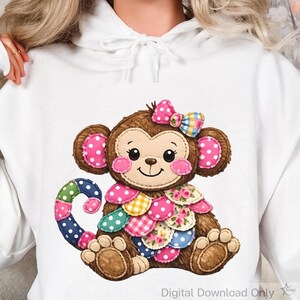 Puede incluir: Una sudadera con capucha blanca con un diseño de mono de dibujos animados. El mono es marrón con orejas de lunares rosas y un lazo a juego. Su vientre está adornado con retazos coloridos y la cola es un arcoíris de lunares. El texto "Digital Download Only" está en la parte inferior.