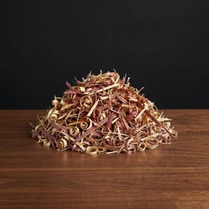 Könnte beinhalten: Ein Haufen aromatischer Zedernholzspäne, der eine Mischung aus hellbraunen und rot-violetten Farbtönen aufweist. Die Holzspäne sind eng gerollt und auf einer dunklen Holzoberfläche mit schwarzem Hintergrund angeordnet.