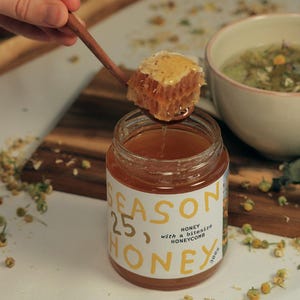 Könnte beinhalten: Ein Honigglas mit der Aufschrift "SEASON 25, HONEY" mit einer mundgerechten Honigwabe. Ein Holzlöffel hält ein Stück Honigwabe, wobei Honig in das Glas tropft. Das Glas ist 300g.