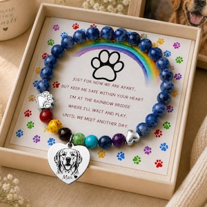 Puede incluir: Una pulsera de cuentas con piedras azules, un dije de huella plateado y un colgante en forma de corazón con el grabado de un perro y el nombre Max. La pulsera se presenta sobre una tarjeta con un arcoíris, huellas y el texto: Just for now we are apart, but keep me safe within your heart I'm at the rainbow bridge where I'll wait and play, until we meet another day.