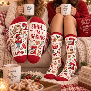 May include: A pair of white crew socks with red cuffs featuring a cartoon illustration of a person in a red apron, a red stand mixer, cupcakes, and the text 'SHHH... I'M BAKING' and 'EMMA' alongside a pattern of baking tools and holiday motifs.
