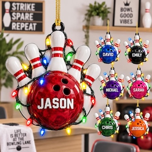 Op de afbeelding: Een gepersonaliseerde kerstversiering met bowlingthema, bestaande uit een rode bowlingbal met de naam JASON in het wit, omringd door witte bowlingkegels en een snoer met gekleurde kerstverlichting, hangend aan een gouden koord.
