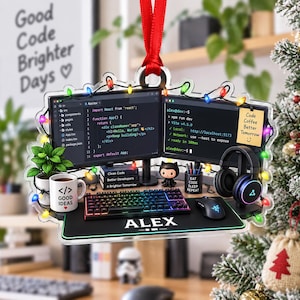 Op de afbeelding: Een doorzichtig acryl ornament met een computeropstelling met twee monitoren, code op de schermen, een mechanisch toetsenbord, muis, een mok met de tekst GOOD IDEAS, een plant, een koptelefoon en een snoer met gekleurde kerstverlichting, gepersonaliseerd met de naam ALEX.