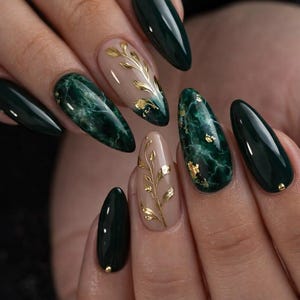 May include: A set of manicured nails featuring a dark green color and marble effect design. Some nails have gold leaf accents and a nude base with gold leaf branch designs. A gold stud is on the base of some nails.