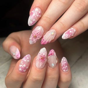 May include: Almond-shaped nails with a professional manicure. The nails are decorated with pink and white floral designs, tiny pearls, and crystals. The overall look is soft and feminine, with a focus on intricate detail.