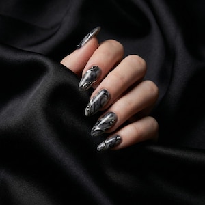 Könnte beinhalten: Eine Hand mit langen, oval geformten Nägeln. Die Nägel weisen ein schwarz-graues Marmormuster mit silbernen Akzenten auf. Die Nägel sind vor einem schwarzen Satinhintergrund platziert, was einen dramatischen Kontrast erzeugt.