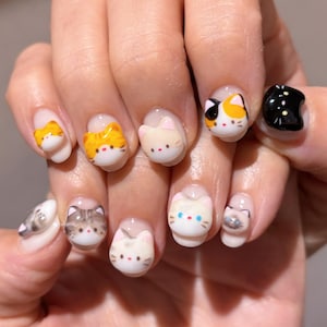Może przedstawiać: Zadbane paznokcie z wzorami kotów 3D. Każdy paznokieć przedstawia inną twarz kota, z różnymi kolorami i wzorami, w tym pomarańczowym, szarym i szylkretowym. Paznokcie mają błyszczące wykończenie.