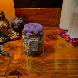 May include: A glass jar filled with dried herbs and sealed with purple wax. A decorative silver charm hangs from the jar. The jar sits on a wooden surface with other spiritual items, including a statue, candles, and crystals.