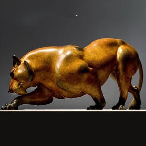 May include: A bronze-toned sculpture of a large predatory cat in a low, crouching pounce position. The figure features a smooth, polished finish with dark shading along the muscular limbs, back, and head, capturing a tense, ready-to-strike posture.