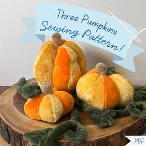 May include: Three plush pumpkins in shades of orange and yellow with brown stems and dark green fabric leaves and vines. A light blue banner at the top displays the text Three Pumpkins Sewing Pattern! and a white needle and thread graphic.