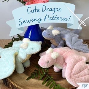 May include: Four soft toy dragons in pastel shades of blue, lilac, and pink. The dragons have wings and are arranged on a wooden base. The image includes the text "Cute Dragon Sewing Pattern!"