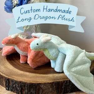 May include: Two plush dragon toys, one in coral pink and one in mint green, featuring soft fabric, small horns, and wings. A light blue banner at the top displays the text Custom Handmade Long Dragon Plush in dark blue cursive lettering.